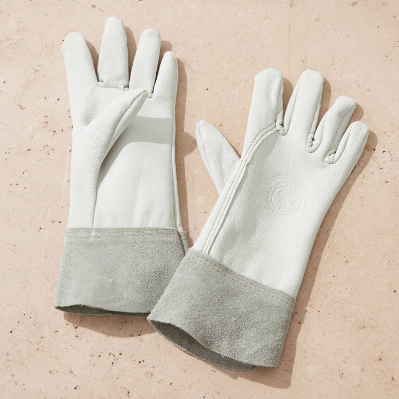 May include: A pair of white leather gardening gloves with grey suede cuffs. The gloves have a stitched seam detail and a small logo on the back of one glove.