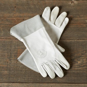 May include: A pair of white leather gloves with a grey cuff. The gloves have a logo on the back of the hand that says "Woman's Work" with a globe and the words "Made & Embroidered in India".