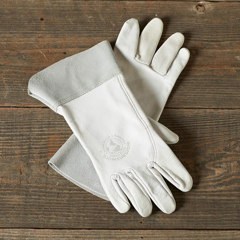 May include: A pair of white leather gloves with a grey cuff. The gloves have a logo on the back of the hand that says "Woman's Work" with a globe and the words "Made & Embroidered in India".