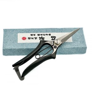 Op de afbeelding: Zwarte tuinscharen met een zilveren mes en een blauwe en witte doos. De doos heeft Japanse tekst die "菊屋 剪定鋏" luidt.
