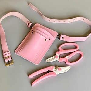 Pu&ograve; includere: Una cintura porta attrezzi da giardinaggio in pelle rosa con una tasca e due cesoie da potatura rosa. Le cesoie hanno la scritta "HAND CRAFTED".