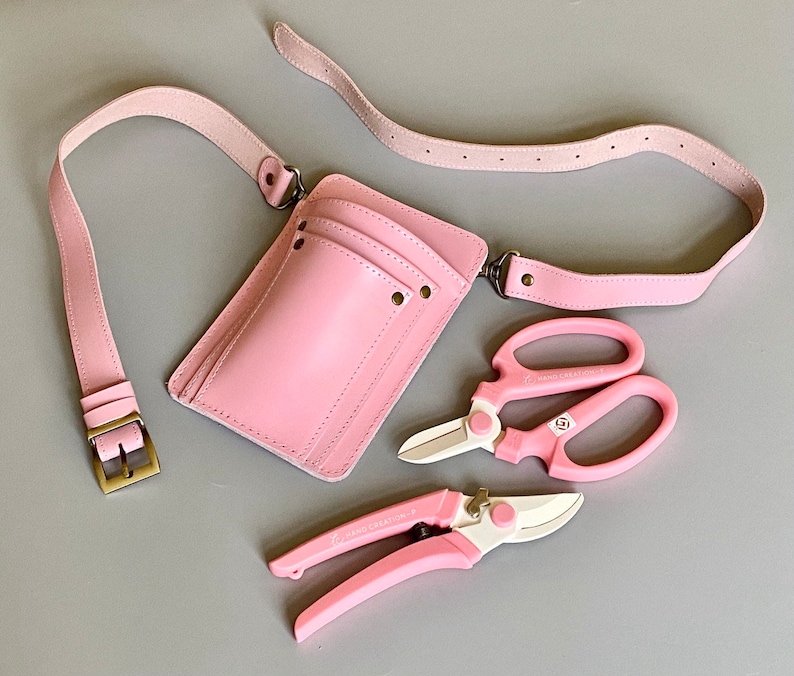 Pu&ograve; includere: Una cintura porta attrezzi da giardinaggio in pelle rosa con una tasca e due cesoie da potatura rosa. Le cesoie hanno la scritta "HAND CRAFTED".