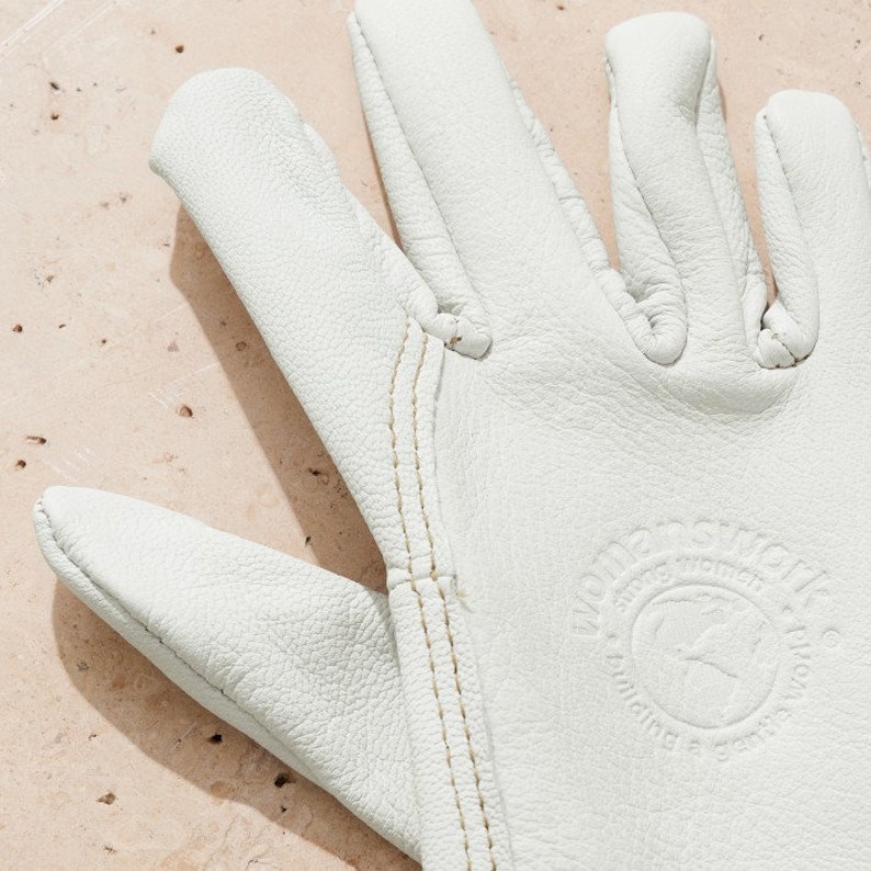 May include: White leather work gloves with a stitched seam down the middle of each finger. The gloves have a circular logo that says "White's  Gloves" and "Handcrafted in the USA".