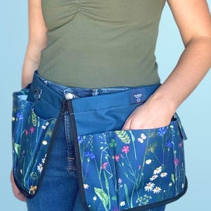 May include: A blue gardening tool belt with multiple pockets and a floral print. The belt is worn over blue jeans and has a black label with the text "Floral Co."
