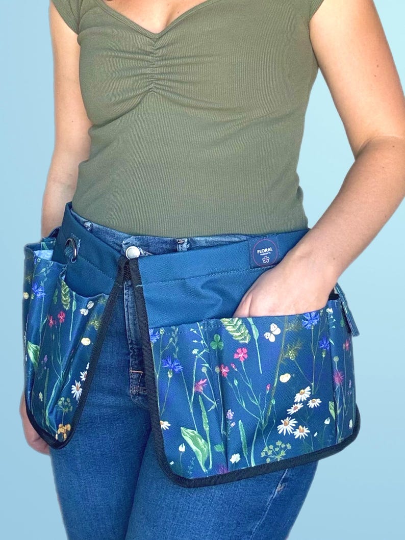 May include: A blue gardening tool belt with multiple pockets and a floral print. The belt is worn over blue jeans and has a black label with the text "Floral Co."