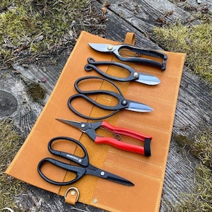 May include: A set of five gardening shears in a brown leather roll. The shears have black and red handles and are arranged in a row. The roll has a flap that closes with a snap.