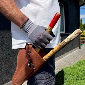 Op de afbeelding: Een bruinleren tuingereedschapsholster met een snoeischaar in de zak. De holster is bevestigd aan de taille van een persoon. De persoon houdt twee tuingereedschappen vast met houten handvatten en rode en gele accenten. De persoon draagt grijze handschoenen.