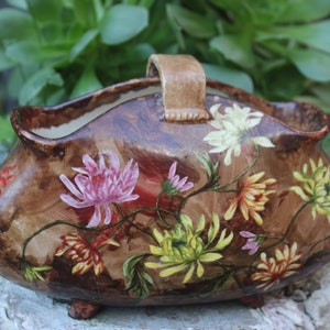 May include: A brown ceramic planter with a handle, decorated with painted flowers in shades of pink, yellow, and red. The planter has three small feet and is shaped like a basket.