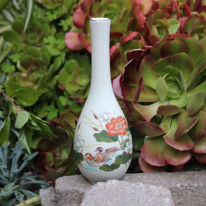 Puede incluir: Un jarr&oacute;n de cer&aacute;mica blanco con un delicado dise&ntilde;o floral y de p&aacute;jaros. El jarr&oacute;n tiene un cuello delgado y un cuerpo redondeado. El dise&ntilde;o presenta una flor roja, flores blancas, hojas verdes y dos p&aacute;jaros.