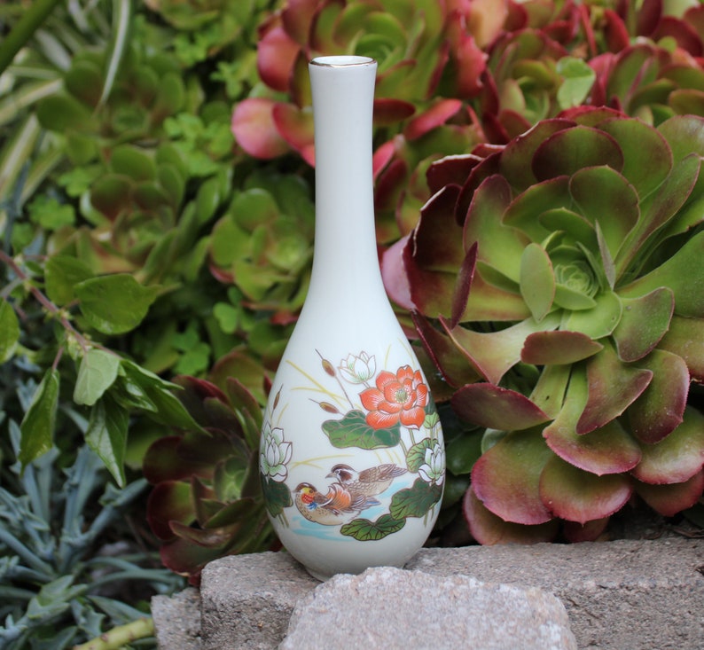 Puede incluir: Un jarr&oacute;n de cer&aacute;mica blanco con un delicado dise&ntilde;o floral y de p&aacute;jaros. El jarr&oacute;n tiene un cuello delgado y un cuerpo redondeado. El dise&ntilde;o presenta una flor roja, flores blancas, hojas verdes y dos p&aacute;jaros.