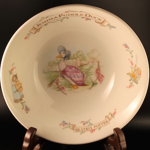 Könnte beinhalten: Eine weiße Keramikschüssel mit einer bunten Illustration von Jemima Puddle-Duck aus Beatrix Potters Kinderbuch. Die Schüssel hat einen blauen und rosa floralen Rand und den Text "Jemima Puddle-Duck" und "Beatrix Potter" in braunen Buchstaben.