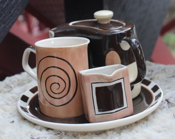 以下が含まれることがあります： 茶色の陶器のティーセットで、ティーポット、マグカップ、クリーマー、ソーサーが付いています。マグカップには茶色の渦巻き模様、クリーマーには茶色の正方形模様、ティーポットには茶色とクリーム色の模様が施されています。