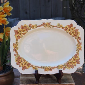 Pode incluir: Uma travessa oval branca para servir com borda ondulada e design floral marrom e amarelo. A travessa está em um suporte de madeira.
