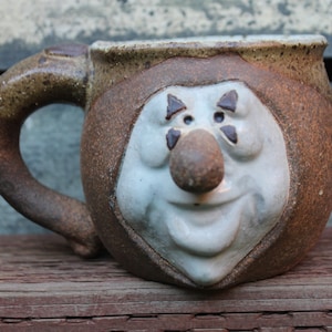 May include: A brown and white ceramic mug with a face sculpted on the front. The face has a large nose, a wide smile, and two black eyes with white triangles.