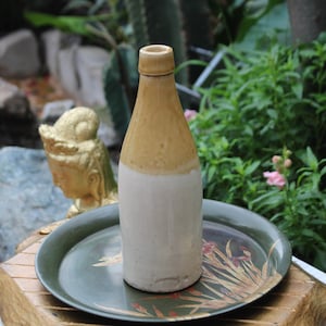 Pode incluir: Uma garrafa de cerâmica com a parte superior marrom e a inferior branca. A garrafa está sobre uma bandeja verde com um design floral.