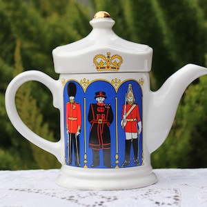 Puede incluir: Tetera de cerámica blanca con un diseño azul y dorado que presenta tres guardias británicos con uniformes rojos y azules. La tetera tiene una tapa con una corona dorada en la parte superior.