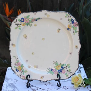 May include: A white square ceramic serving plate with a black rim and a floral design in shades of pink, yellow, blue, and green.