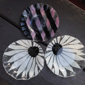 Puede incluir: Tres platos decorativos de vidrio con diferentes diseños. Un plato tiene un patrón geométrico negro y rosa, otro tiene un diseño de margarita blanca y el tercero tiene un diseño de margarita blanco y negro.