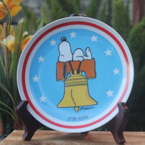 May include: A blue and white ceramic plate with a red and white border. The plate features a cartoon image of Snoopy laying on top of the Liberty Bell. The plate has stars around the border and the text "Schultz" and "1776-1976" at the bottom.