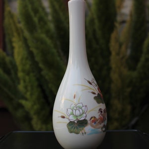 Puede incluir: Jarr&oacute;n de cer&aacute;mica blanca con un dise&ntilde;o floral que presenta una flor de loto y dos p&aacute;jaros. El jarr&oacute;n tiene un cuello estrecho y un cuerpo redondeado.