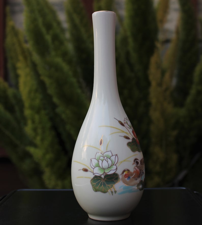 Puede incluir: Jarr&oacute;n de cer&aacute;mica blanca con un dise&ntilde;o floral que presenta una flor de loto y dos p&aacute;jaros. El jarr&oacute;n tiene un cuello estrecho y un cuerpo redondeado.