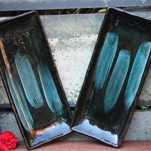 May include: Two rectangular ceramic serving dishes with black glaze and teal blue brushstrokes. The dishes are sitting on a wooden surface.