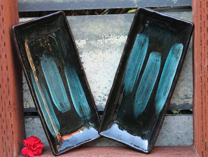 May include: Two rectangular ceramic serving dishes with black glaze and teal blue brushstrokes. The dishes are sitting on a wooden surface.