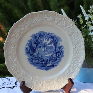 May include: A white ceramic plate with a blue and white floral design. The plate has a raised rim and a scalloped edge. The design features a blue and white floral pattern with a central image of a castle in a landscape.