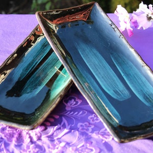 May include: Two rectangular ceramic serving dishes with a dark blue glaze and a light green brushstroke design. The dishes are stacked on top of each other and are sitting on a purple fabric surface.