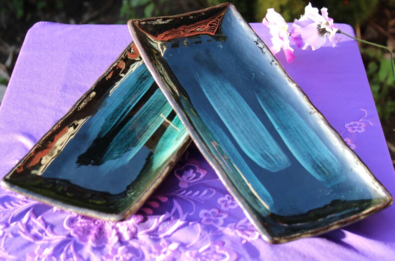 May include: Two rectangular ceramic serving dishes with a dark blue glaze and a light green brushstroke design. The dishes are stacked on top of each other and are sitting on a purple fabric surface.