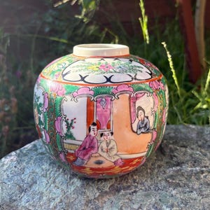 Puede incluir: Un tarro de jengibre redondo de porcelana con escenas pintadas a mano de figuras con vestimenta tradicional dentro de paneles arqueados. El recipiente está decorado con motivos florales vibrantes en rosa, verde y naranja, detalles dorados.