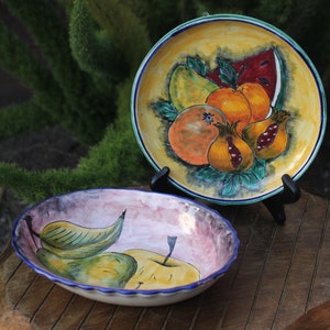 Könnte beinhalten: Zwei Keramikteller mit einem Fruchtdesign. Der größere Teller hat einen gelben Hintergrund mit einer Wassermelone, Birnen, Orangen und Granatäpfeln. Der kleinere Teller hat einen rosa Hintergrund mit Birnen und Äpfeln.
