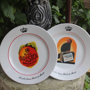 May include: Two white ceramic plates with red trim and gold crowns. One plate features a peach with the text "Hotel Bristol Barcelona" and the phrase "Les plus beaux Hotels du Monde". The other plate features a black bird with a suitcase and the text "DGARD MONTEVIDEO" and the phrase "plus beaux Hotels du Monde".