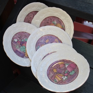 May include: Set of five white ceramic plates with a purple and pink floral design and gold accents. The plates feature a variety of fruits and vegetables, including grapes, apples, pears, and plums. The text "La Surpresa" is printed on the bottom of one plate.