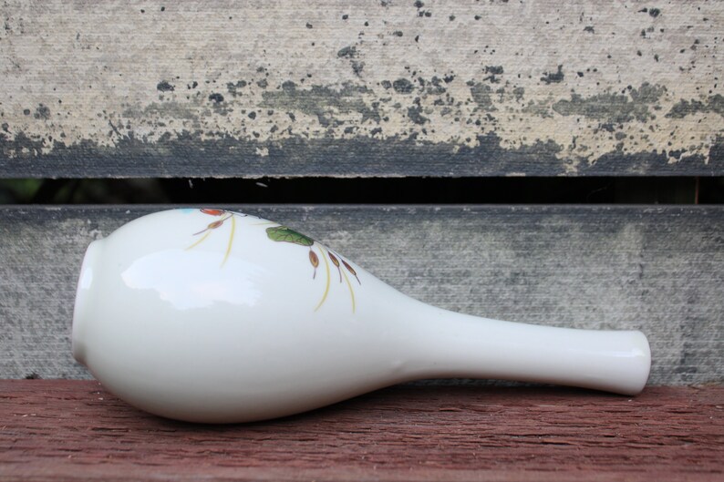 Puede incluir: Jarr&oacute;n de cer&aacute;mica blanco con un dise&ntilde;o floral en tonos rojos, verdes y amarillos. El jarr&oacute;n tiene un cuello largo y delgado y un cuerpo redondeado.