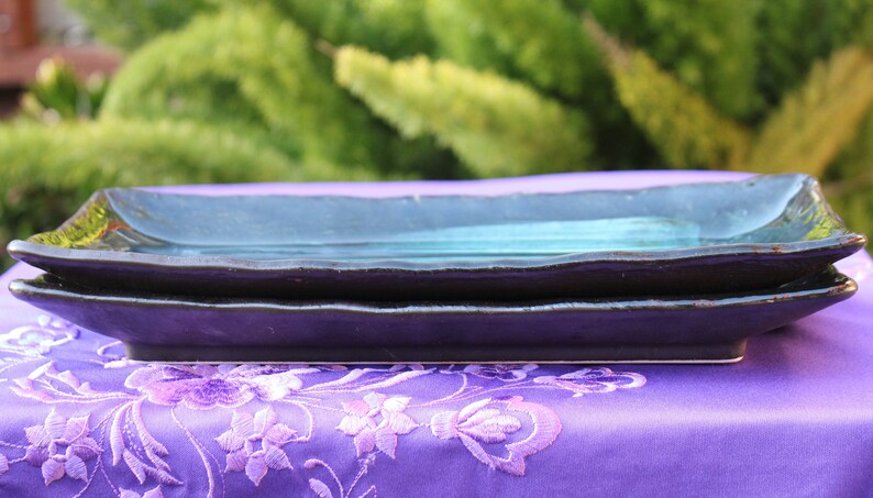 May include: Two rectangular ceramic serving plates stacked on top of each other. The top plate is a dark teal color and the bottom plate is a dark purple color. Both plates have a slightly wavy edge.