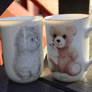 May include: Two white ceramic mugs with gold trim. One mug features a gray kitten with blue eyes. The other mug features a brown teddy bear holding a white kitten. Both mugs have a gold rim.