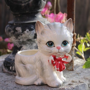 May include: White ceramic cat planter with a red bow. The cat has gray markings and blue eyes. The planter has a small opening at the top for planting.