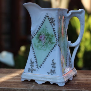 May include: A white porcelain pitcher with a diamond-shaped design and floral pattern. The pitcher has a gold trim and a handle.