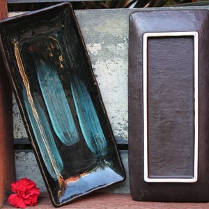 May include: Two rectangular ceramic serving dishes. One dish is black with teal blue streaks and gold accents. The other dish is brown with a white border.