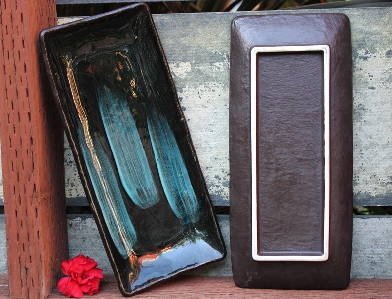 May include: Two rectangular ceramic serving dishes. One dish is black with teal blue streaks and gold accents. The other dish is brown with a white border.