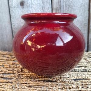 May include: A small, round, glossy ceramic pot with a deep red finish. The vessel features a wide, bulbous body that tapers towards a smooth, circular opening at the top. The surface is highly reflective, showing light highlights across the curve.