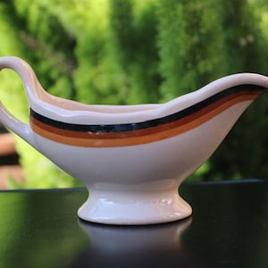 Może przedstawiać: Biała ceramiczna sosjerka z brązowo-czarno-ciemnoniebieskim paskowanym wzorem. Sosjerka ma uchwyt i podstawę.
