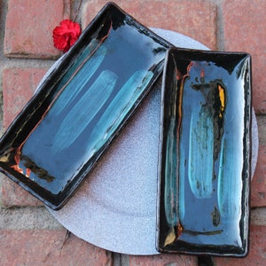 May include: Two rectangular ceramic serving dishes with black glaze and teal blue brushstrokes. The dishes are on a gray background.