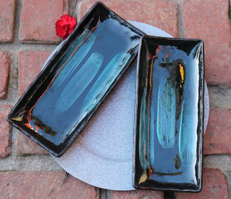 May include: Two rectangular ceramic serving dishes with black glaze and teal blue brushstrokes. The dishes are on a gray background.