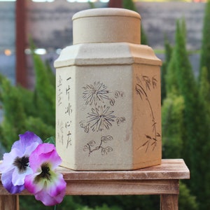 May include: A beige ceramic tea caddy with a hexagonal shape and a lid. The caddy is decorated with a floral design and Chinese characters. The caddy is sitting on a wooden table.