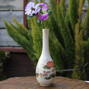 Puede incluir: Un jarr&oacute;n de cer&aacute;mica blanco con un dise&ntilde;o floral que presenta p&aacute;jaros y flores. El jarr&oacute;n tiene un cuello largo y delgado y una base redondeada. Est&aacute; lleno con un solo tallo de flores moradas y blancas.