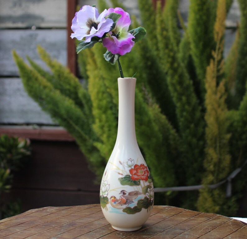 Puede incluir: Un jarr&oacute;n de cer&aacute;mica blanco con un dise&ntilde;o floral que presenta p&aacute;jaros y flores. El jarr&oacute;n tiene un cuello largo y delgado y una base redondeada. Est&aacute; lleno con un solo tallo de flores moradas y blancas.