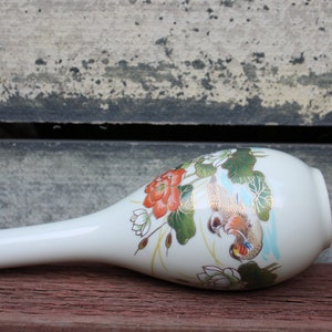 Puede incluir: Un jarr&oacute;n de cer&aacute;mica blanca con un cuello largo y delgado y un cuerpo redondeado. El jarr&oacute;n est&aacute; decorado con un dise&ntilde;o floral con flores rojas y blancas, hojas verdes y detalles dorados. Dos p&aacute;jaros est&aacute;n representados en el jarr&oacute;n, uno posado en una rama y el otro en vuelo.