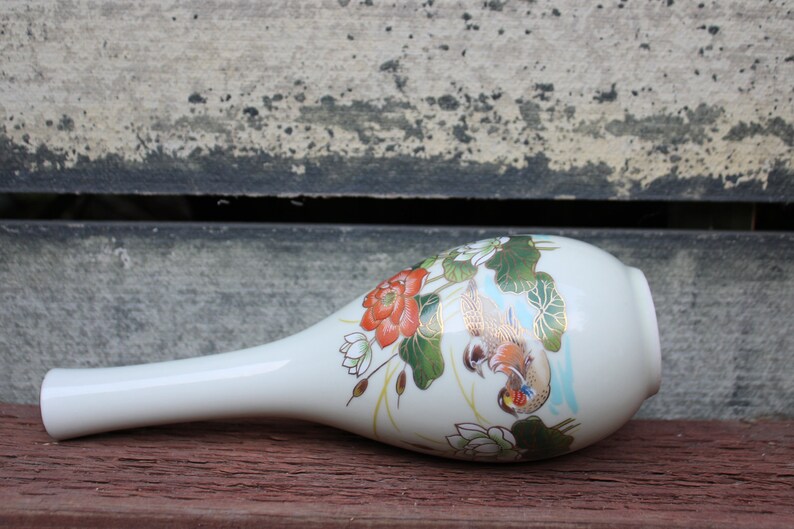 Puede incluir: Un jarr&oacute;n de cer&aacute;mica blanca con un cuello largo y delgado y un cuerpo redondeado. El jarr&oacute;n est&aacute; decorado con un dise&ntilde;o floral con flores rojas y blancas, hojas verdes y detalles dorados. Dos p&aacute;jaros est&aacute;n representados en el jarr&oacute;n, uno posado en una rama y el otro en vuelo.